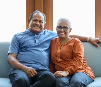 Smiling older couple sitting together on a couch at home, arm-in-arm, looking at the camera.